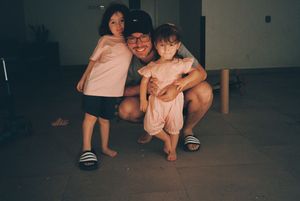 A man crouching and smiling with two young girls in a dimly lit indoor space, all wearing casual summer clothing.