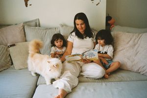 Woman reading a book on a sofa with two young children and a fluffy white cat in a cozy living room.