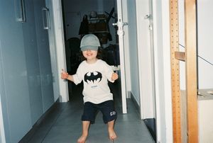 Young child wearing a Batman shirt and gray cap standing barefoot in a hallway, arms outstretched.