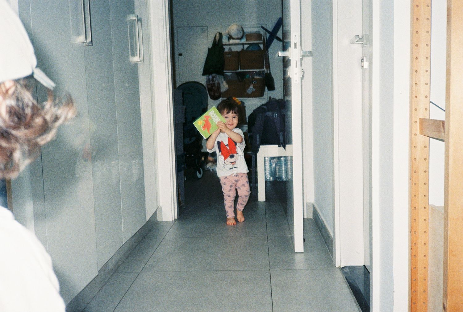 Young toddler walking down a hallway holding a colorful book, wearing pink patterned pajamas in a home setting.