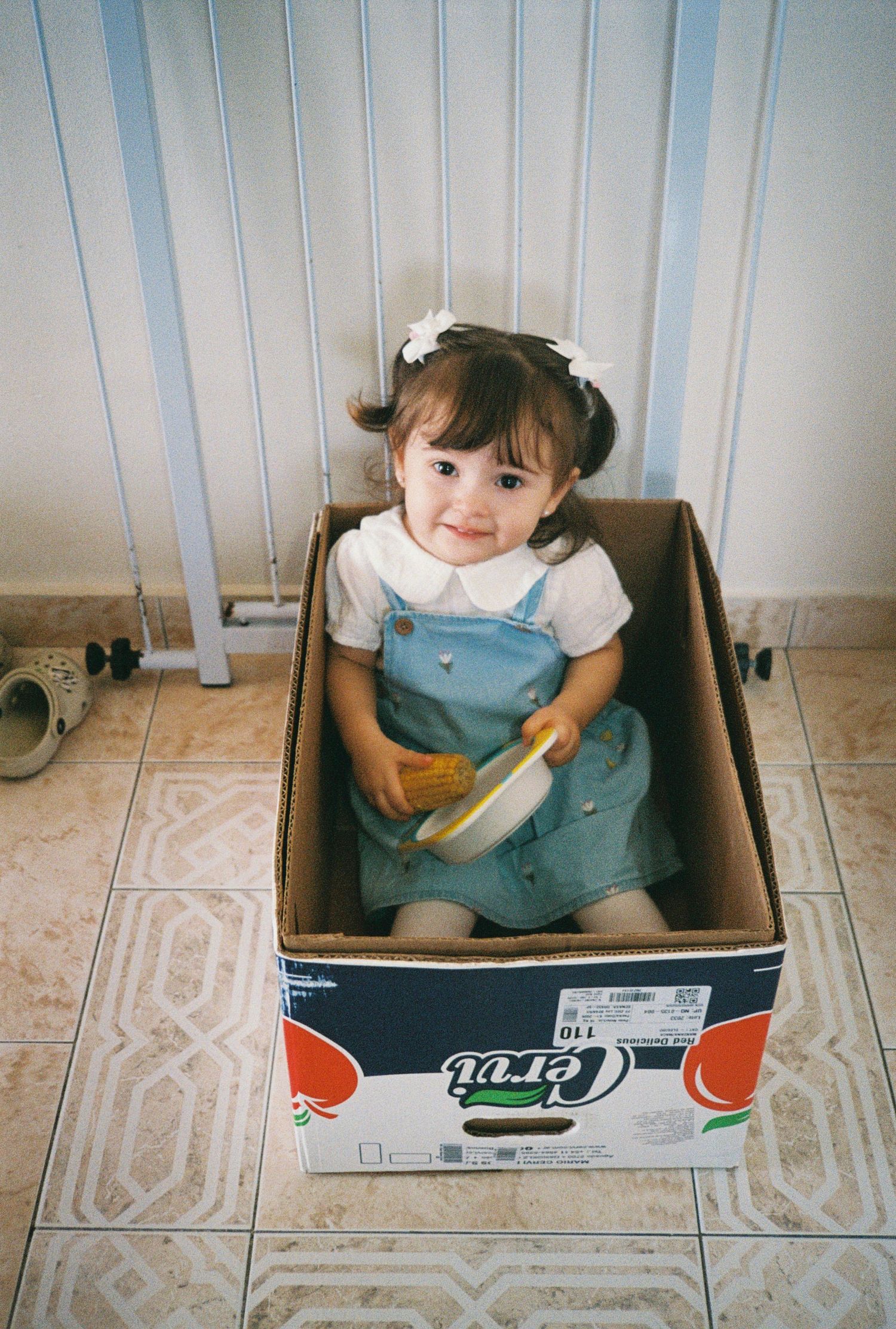 A smiling toddler girl with pigtails sits inside a cardboard box on a tiled floor, holding a yellow toy.
