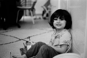 A smiling young child with a bowl cut sits against a wall holding a toy, in a vintage black and white photo.