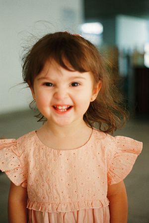 Smiling toddler girl with brown hair wearing a pink eyelet dress, cheerful expression, indoor portrait photo.