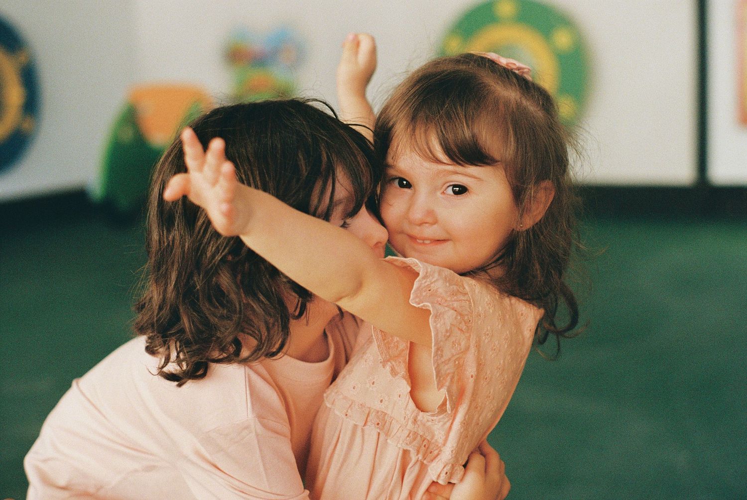 Two young girls in pink dresses hugging and playing together in a colorful classroom setting.