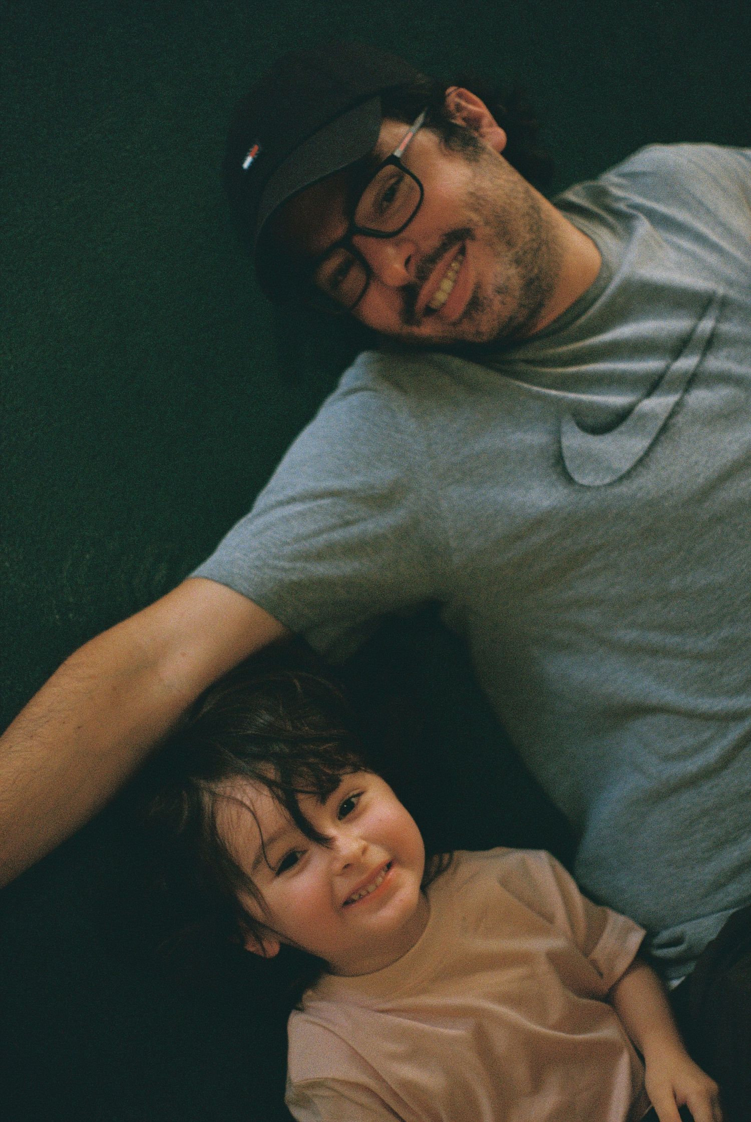 A smiling man wearing glasses and a Nike shirt lies beside a happy toddler on a dark floor.