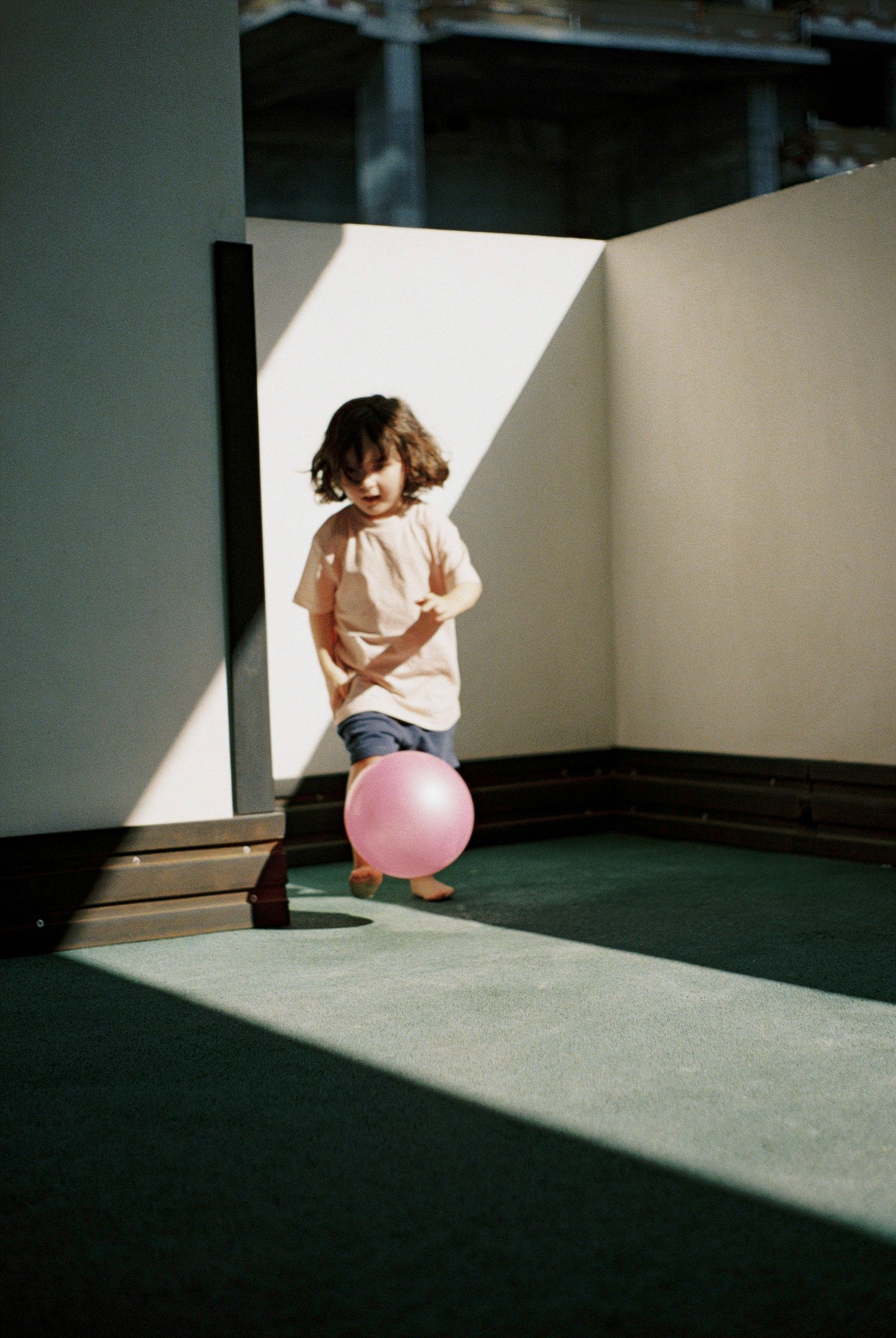 A young child plays with a pink balloon on a green carpet floor in a sunlit corner with white walls.