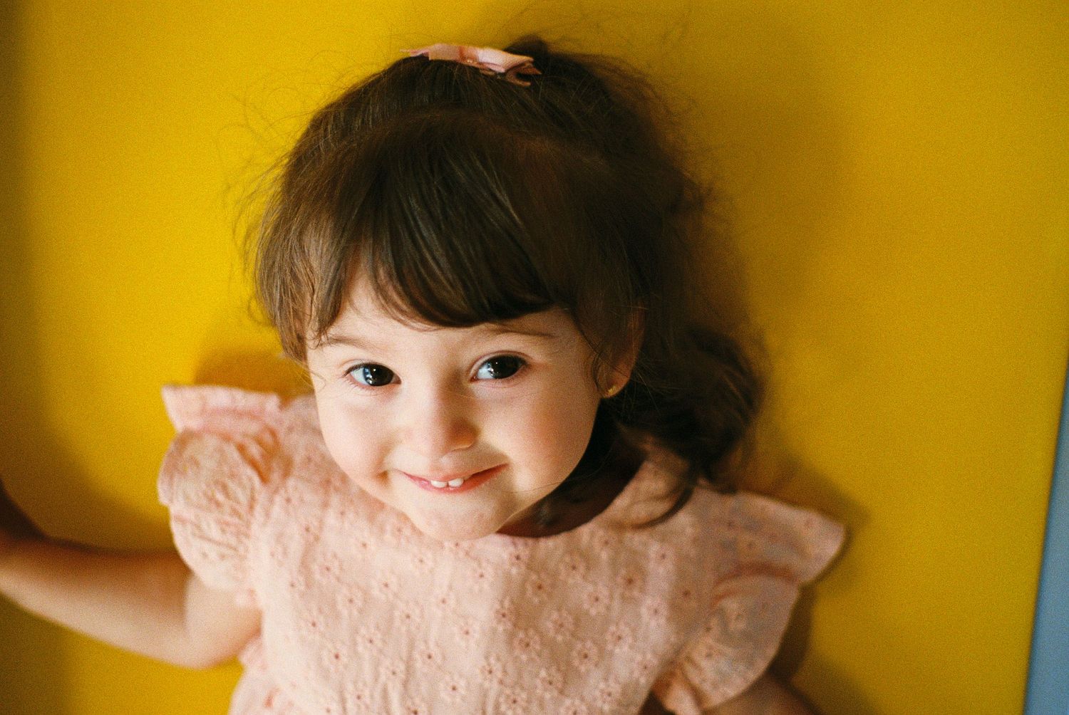 Smiling toddler girl with dark hair and pink bow wearing a pink dress against a bright yellow background.