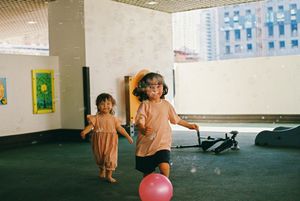 Two young girls playing with a pink balloon indoors near large windows in a bright modern space.