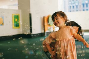 A joyful young girl in a pink dress plays among floating soap bubbles in a colorful indoor play area.