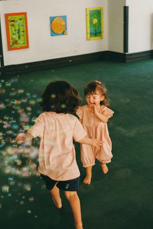 Two young children in pink outfits joyfully chasing bubbles on a green carpet in a colorful art-filled room.