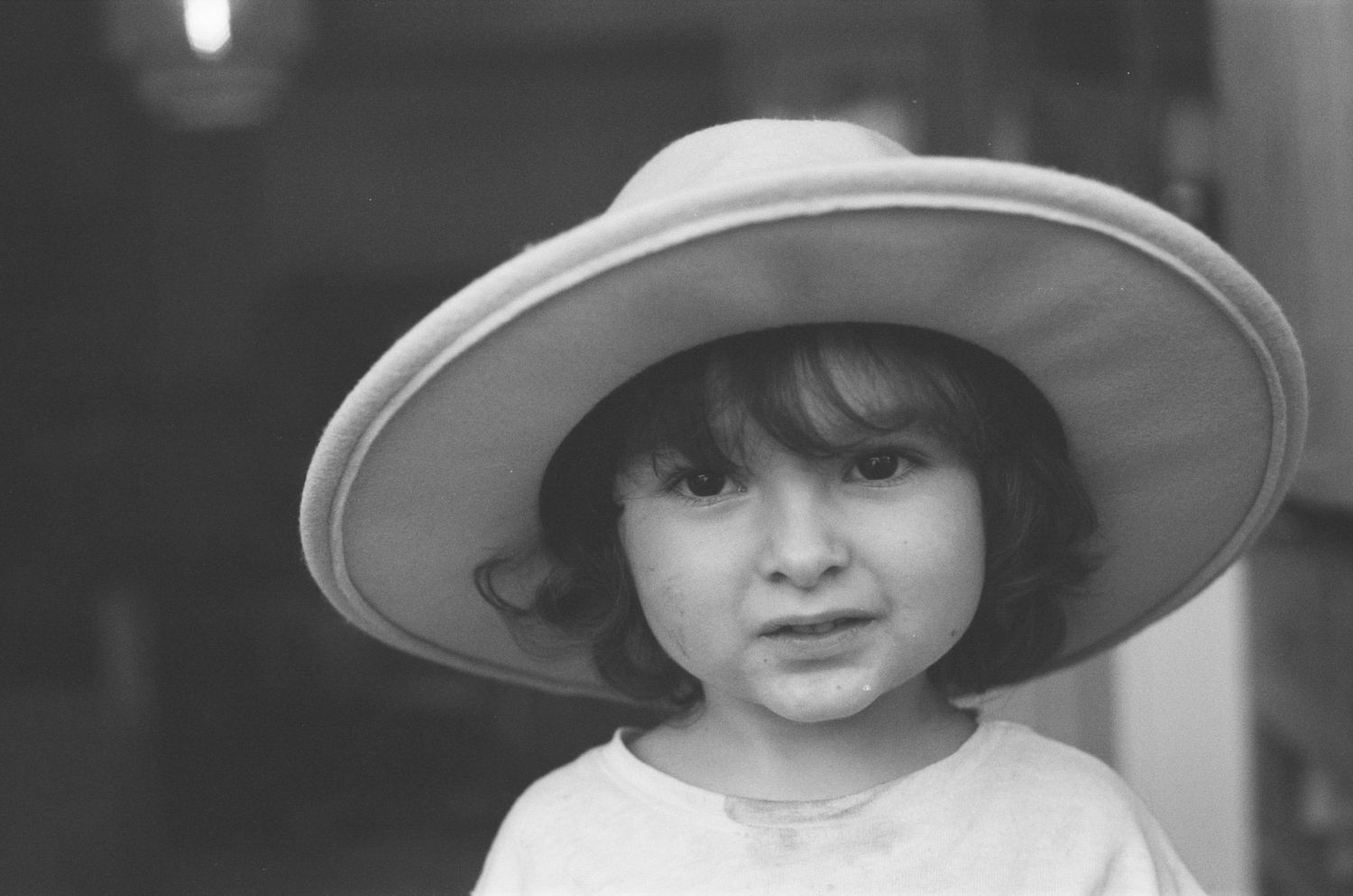 Black and white photo of a young girl wearing an oversized wide-brim hat, looking at the camera.