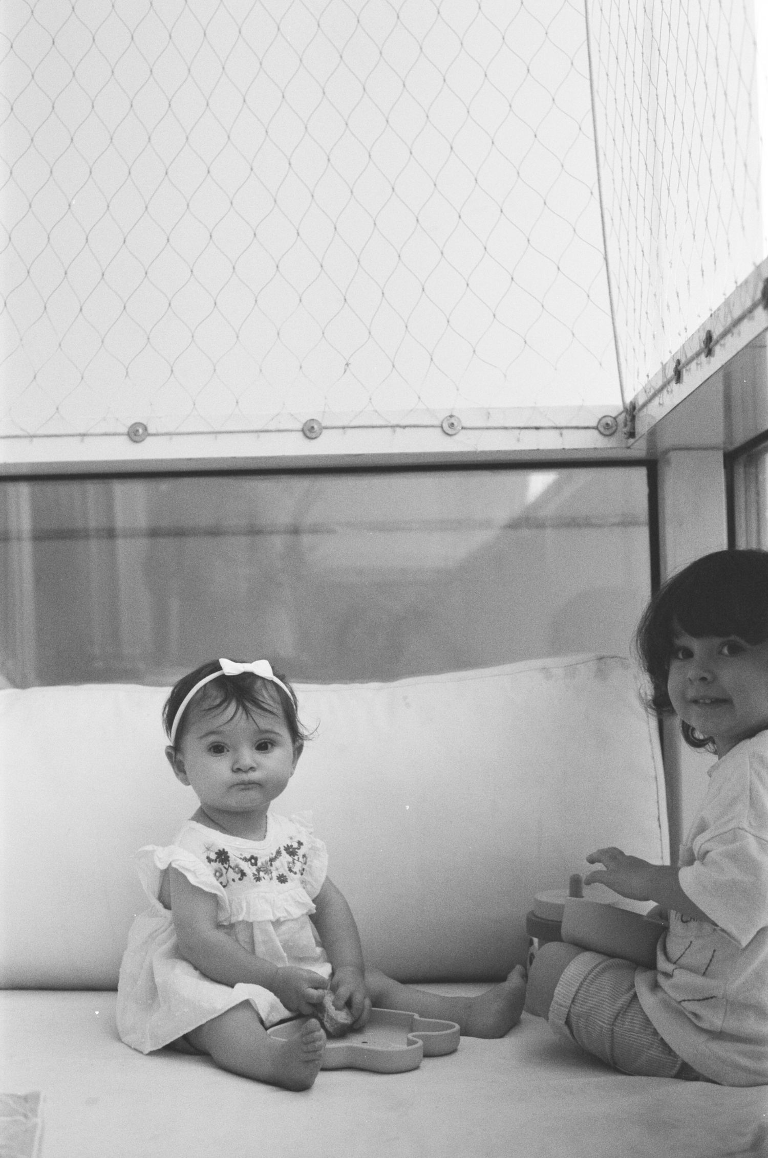 Black and white photo of two young children sitting on a bench, a baby girl in a white dress with a bow headband.