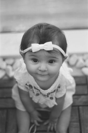 Black and white portrait of an adorable baby girl wearing a white bow headband and lace dress, looking up at camera.