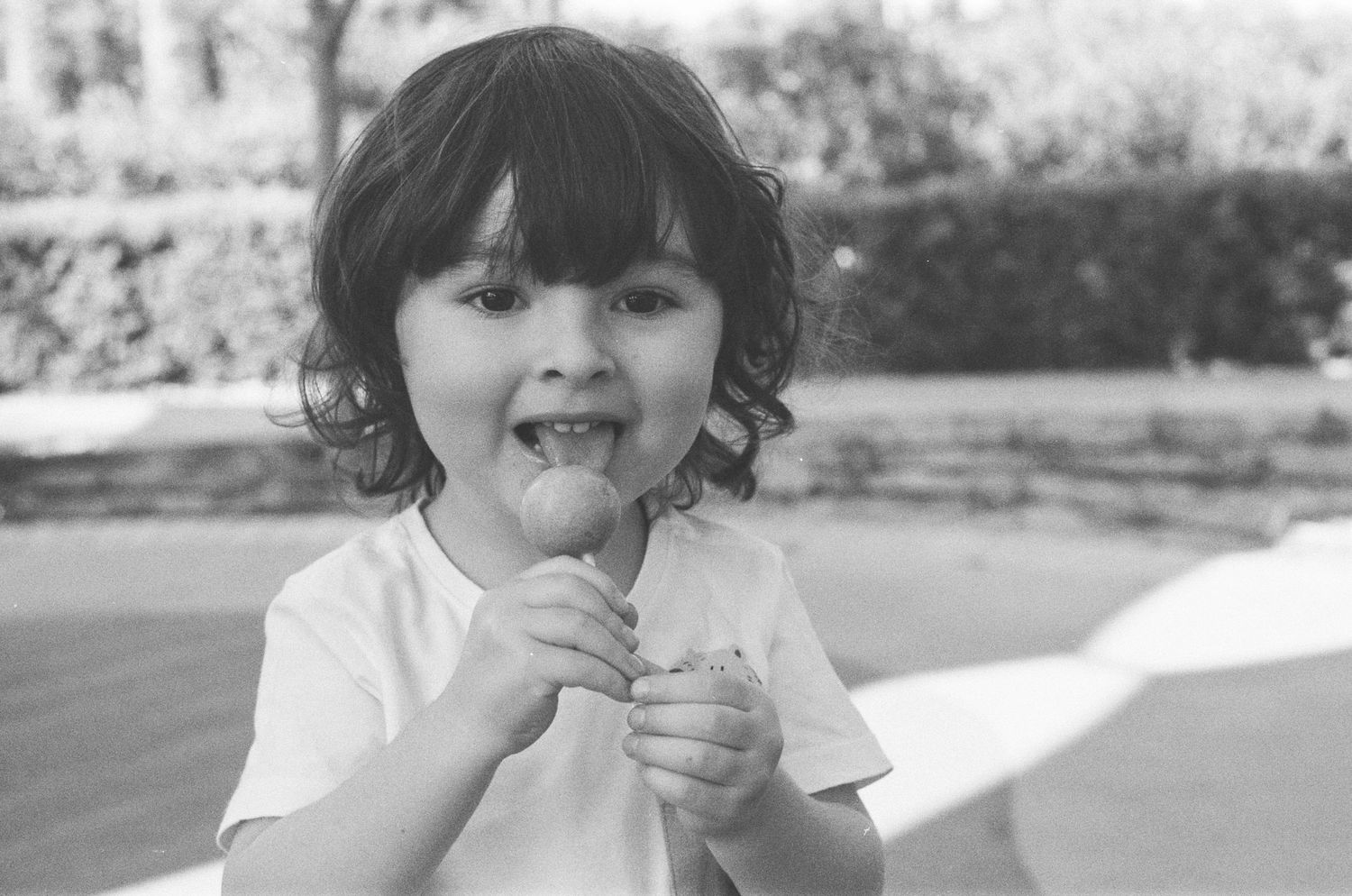 Black and white photo of a happy young child eating a lollipop outdoors in a white t-shirt.