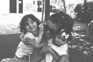 Black and white photo of a smiling adult in sunglasses hugging two young children outdoors.