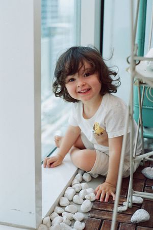 A smiling young child with curly hair sits on a balcony floor surrounded by white pebbles, wearing a white outfit.