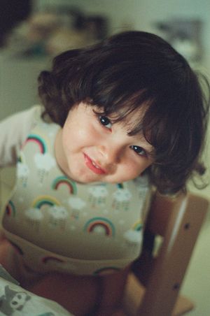 A smiling toddler with curly dark hair wearing a colorful rainbow-print bib sits in a wooden high chair.