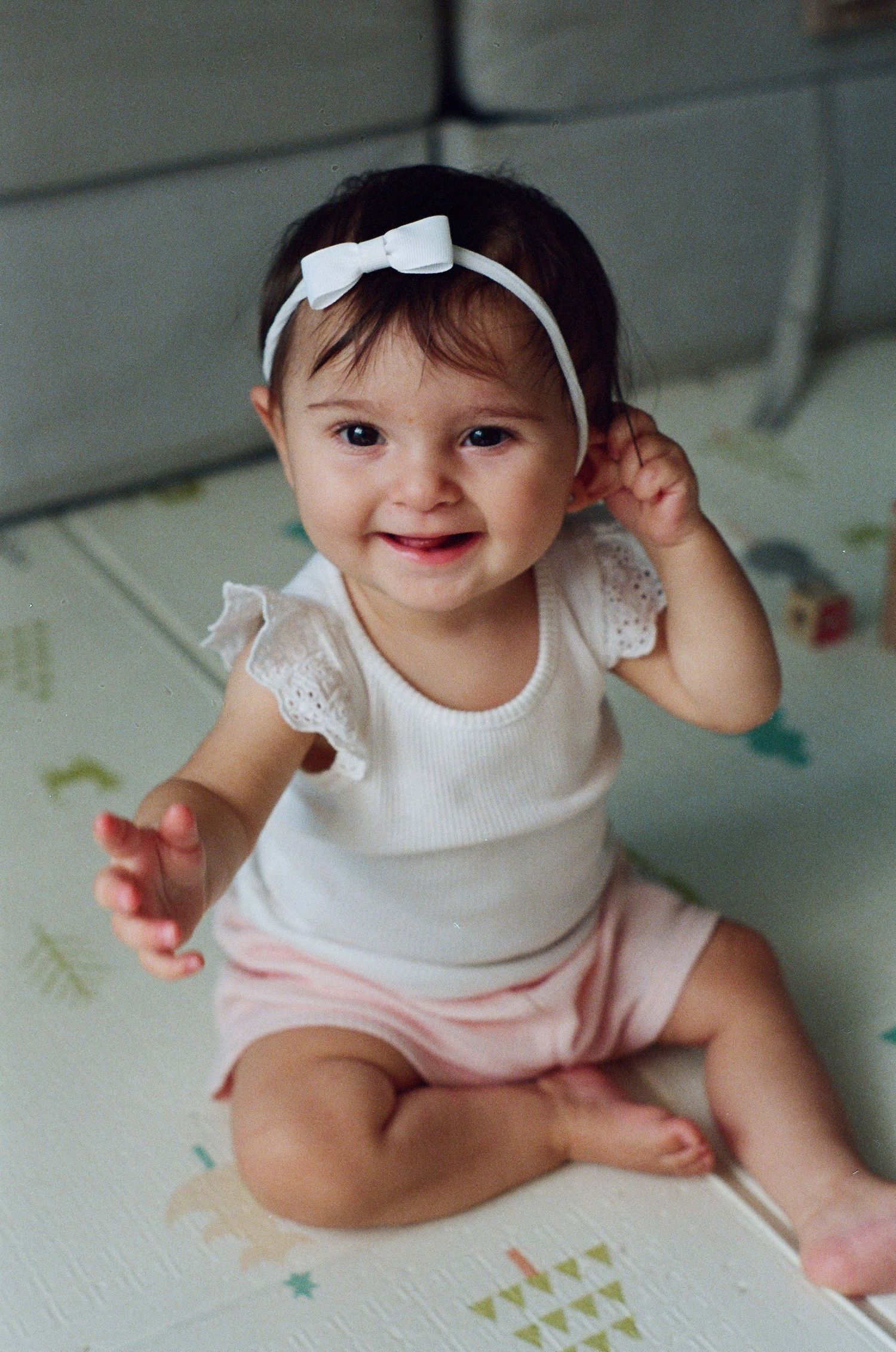 Smiling baby girl in white ruffled top and pink shorts wearing a white bow headband sitting on a play mat.