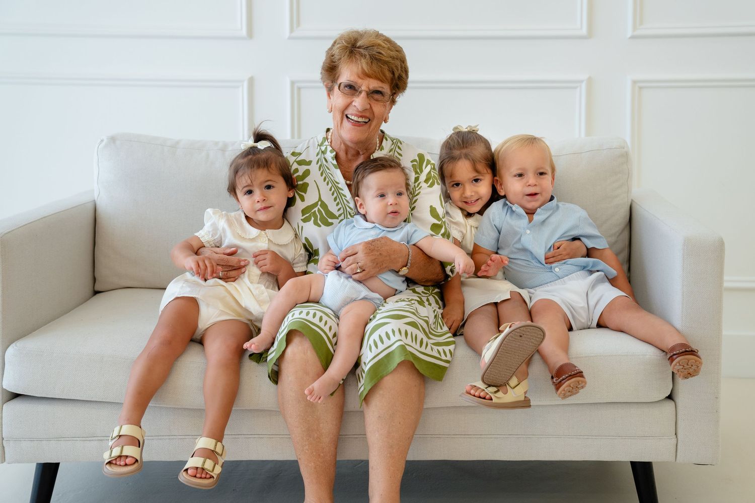 Grandmother sitting on white sofa smiling with four young grandchildren in coordinating outfits.