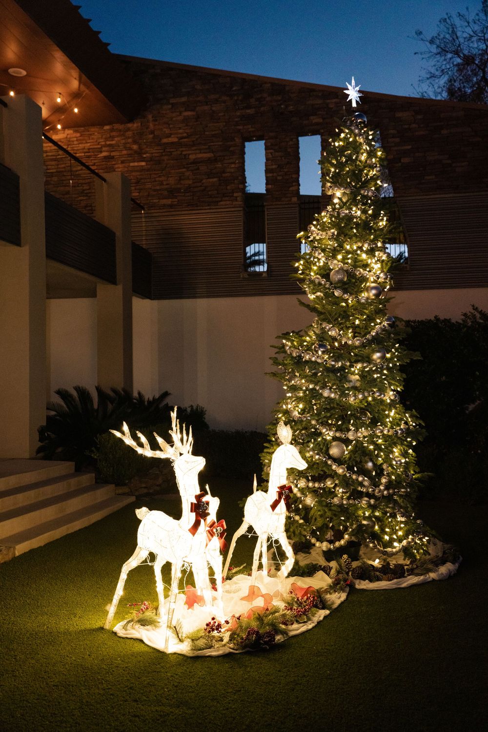 Illuminated reindeer decorations beside a decorated Christmas tree with lights and star topper outside a modern home.
