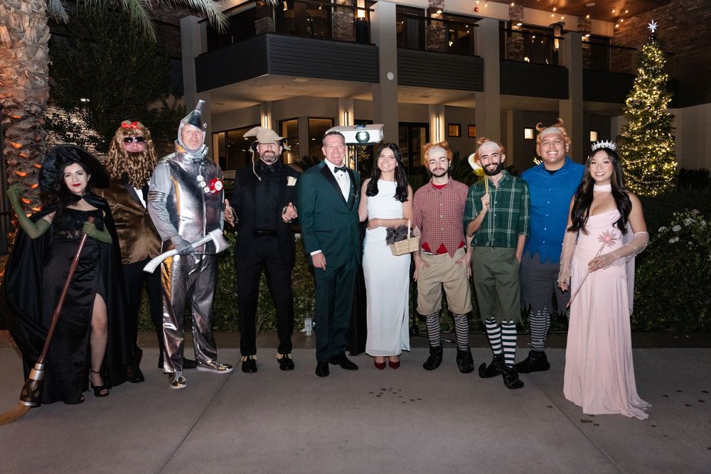 Group of adults in Halloween costumes posing together outdoors at a festive evening party near holiday decorations.