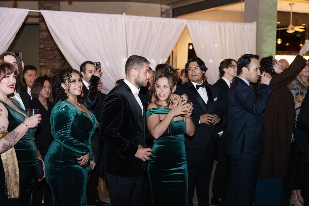 Guests in elegant green velvet and black tuxedo attire mingle and dance at a formal wedding reception.