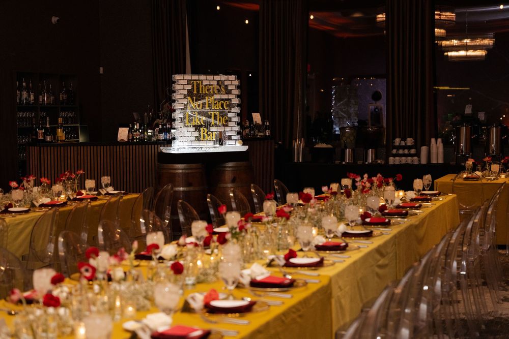 Elegant banquet hall with long gold-draped tables, red floral centerpieces, crystal glassware, and a glowing bar backdrop.