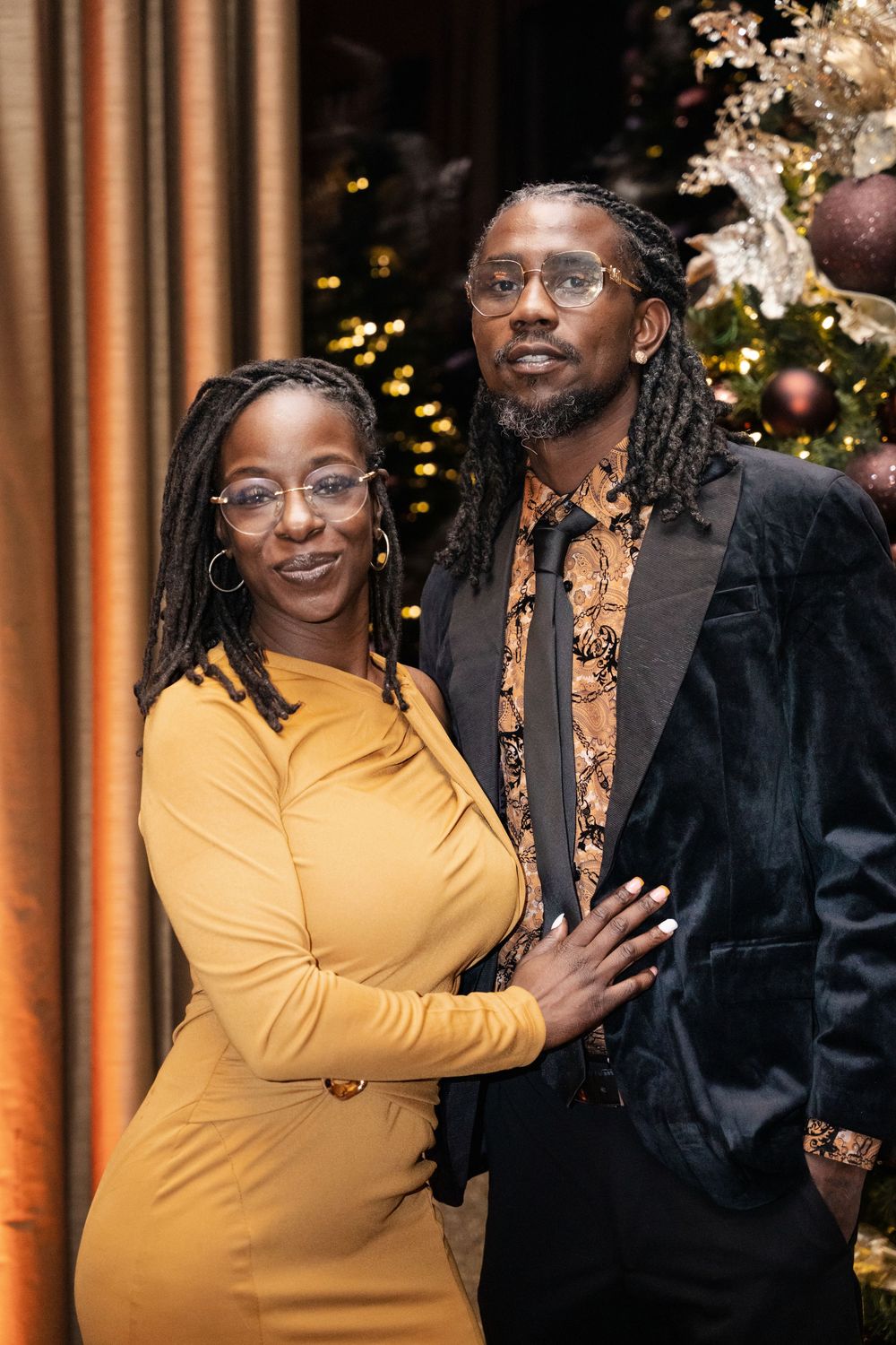 Couple in elegant attire posing together in front of a decorated Christmas tree at a festive event.