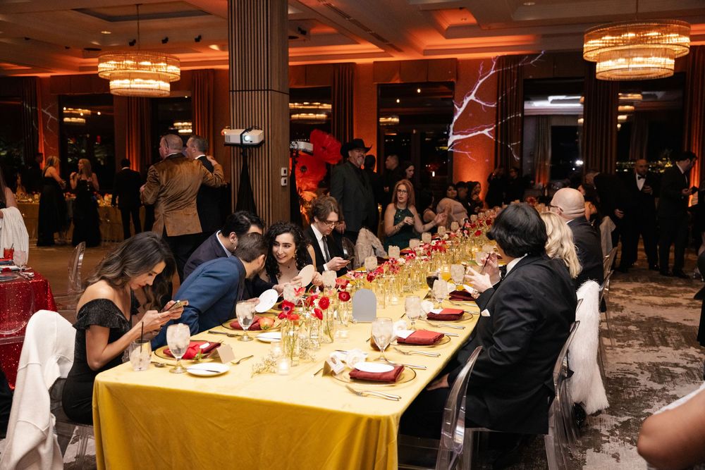 Elegant gala dinner event with guests seated at gold-draped tables adorned with red floral centerpieces in a grand ballroom.