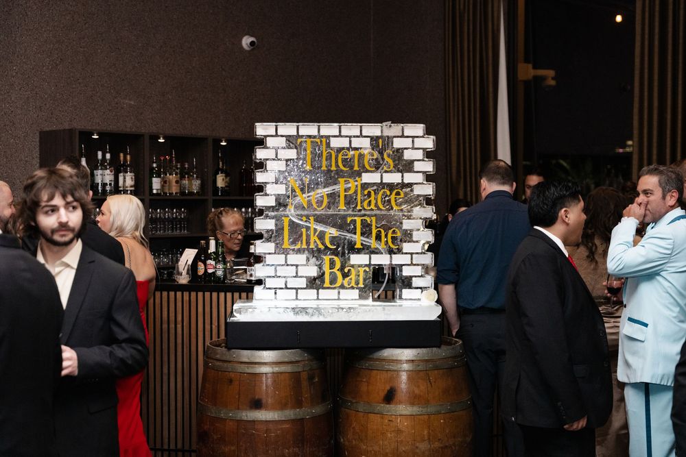 Elegant bar setup with barrel tables and a decorative sign reading There No Place Like The Bar at a formal event.