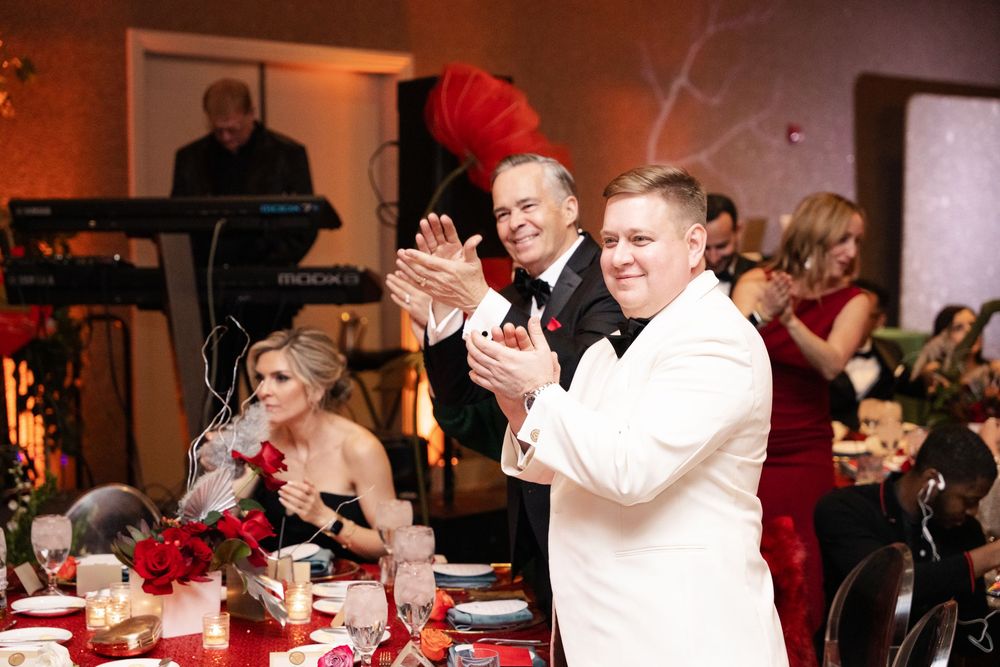 Guests in formal attire clapping at an elegant wedding reception with red floral centerpieces and warm ambient lighting.