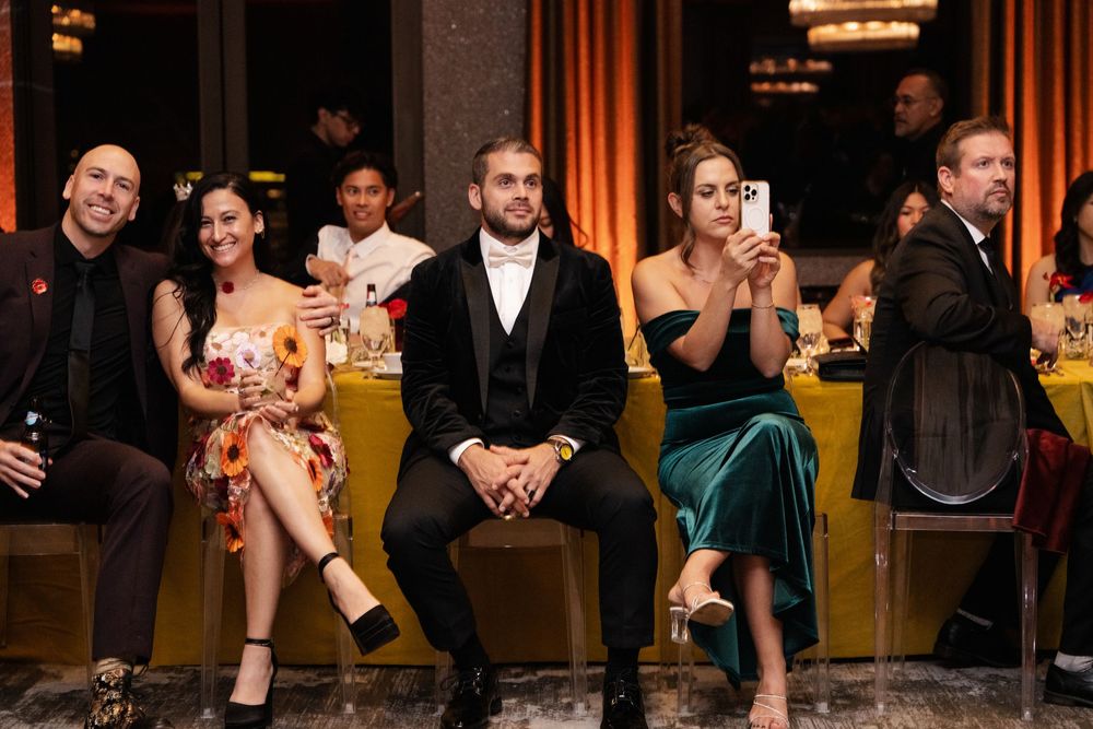 Elegantly dressed guests seated at a formal event, smiling and taking photos in a warmly lit venue.