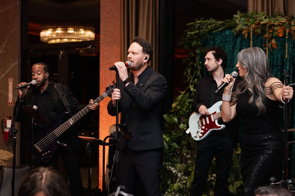 Live band performing on stage in elegant venue, featuring vocalist, guitarist, and bassist in formal black attire.