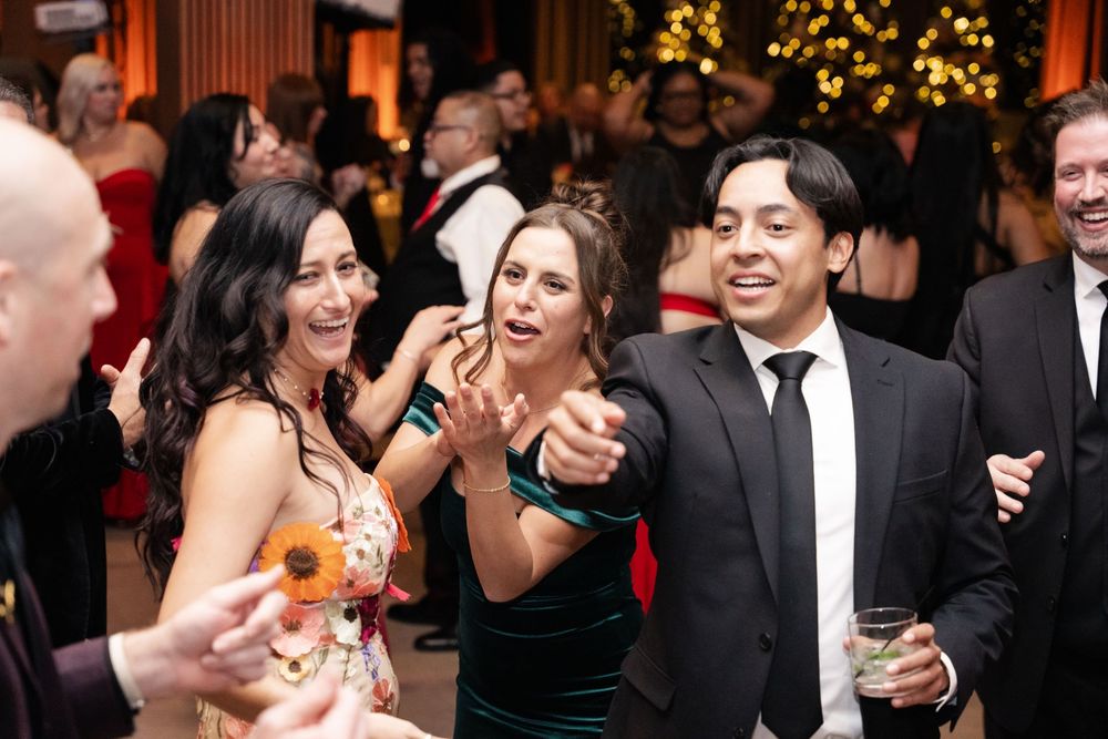 Guests in formal attire laugh and dance together at an elegant holiday gala event with festive decorations.