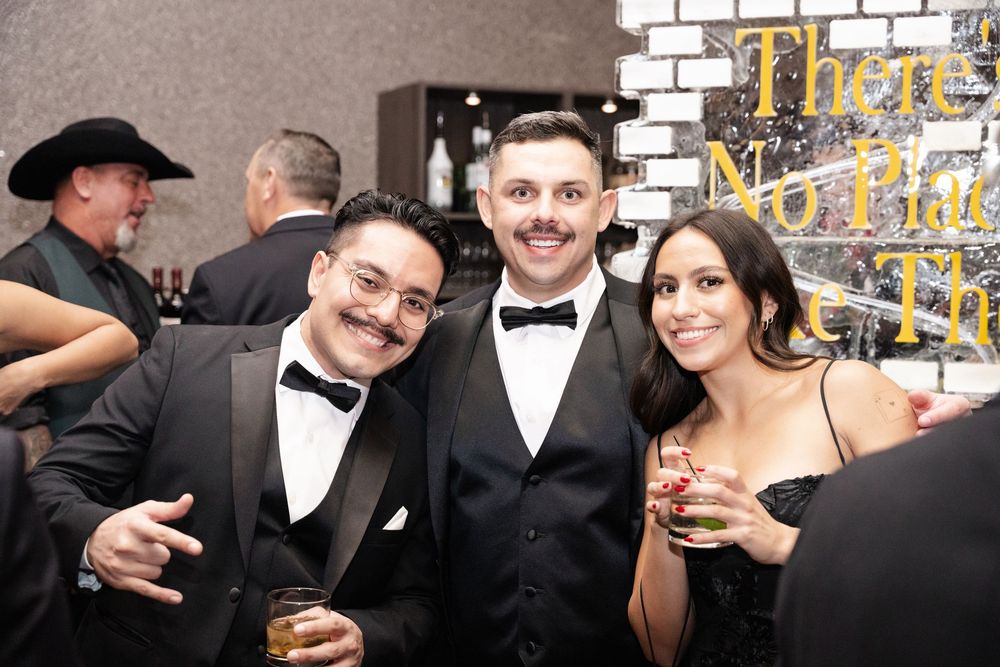 Three guests in formal attire smiling at an elegant black-tie event, holding drinks near a decorative wall.