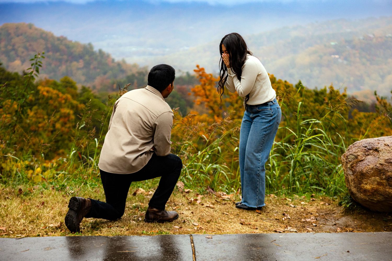Man proposing on one knee to woman at scenic mountain overlook during autumn, with colorful fall foliage in background.
