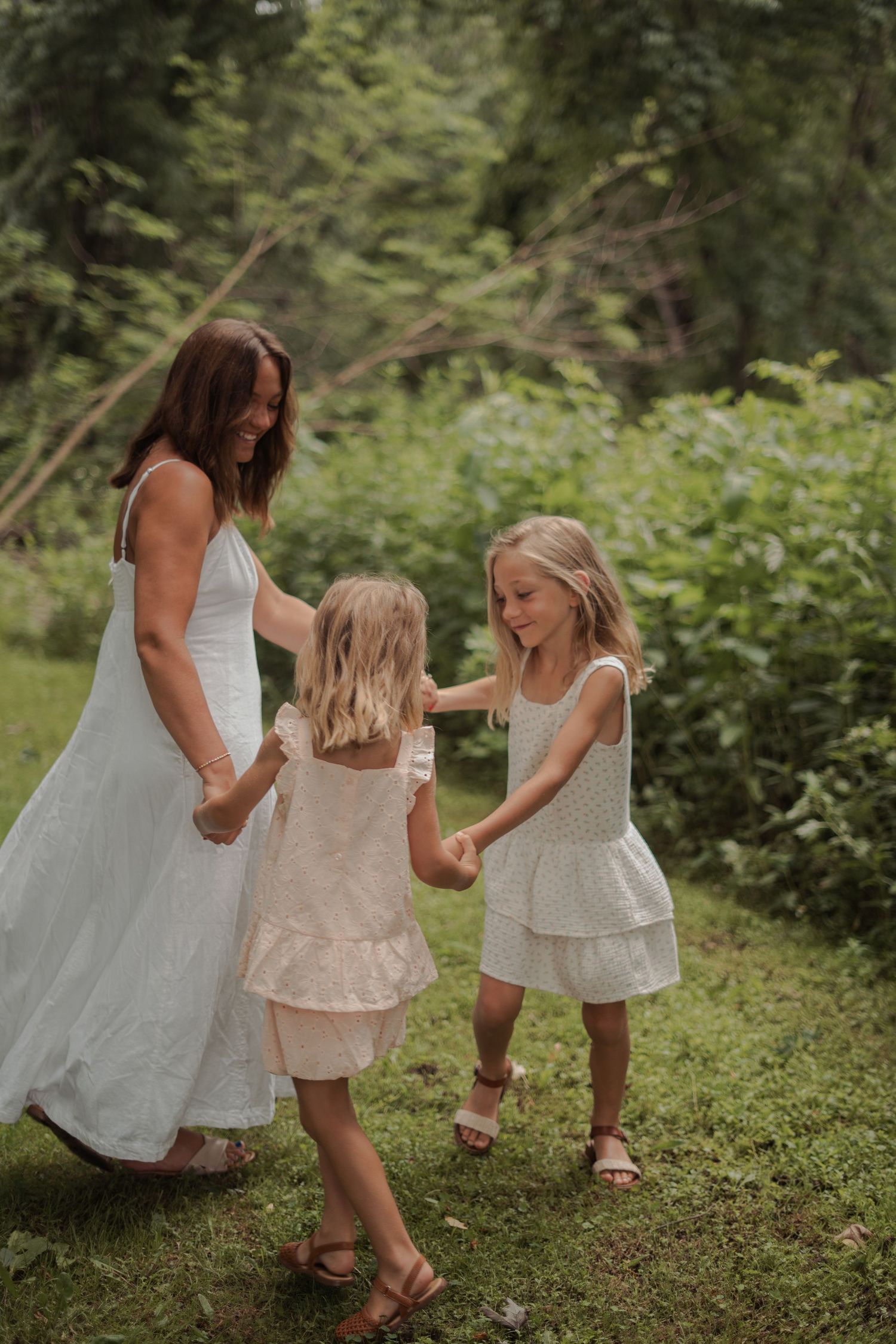 A mother in a white dress holds hands with her two young daughters in white outfits, dancing outdoors in a lush green setting.