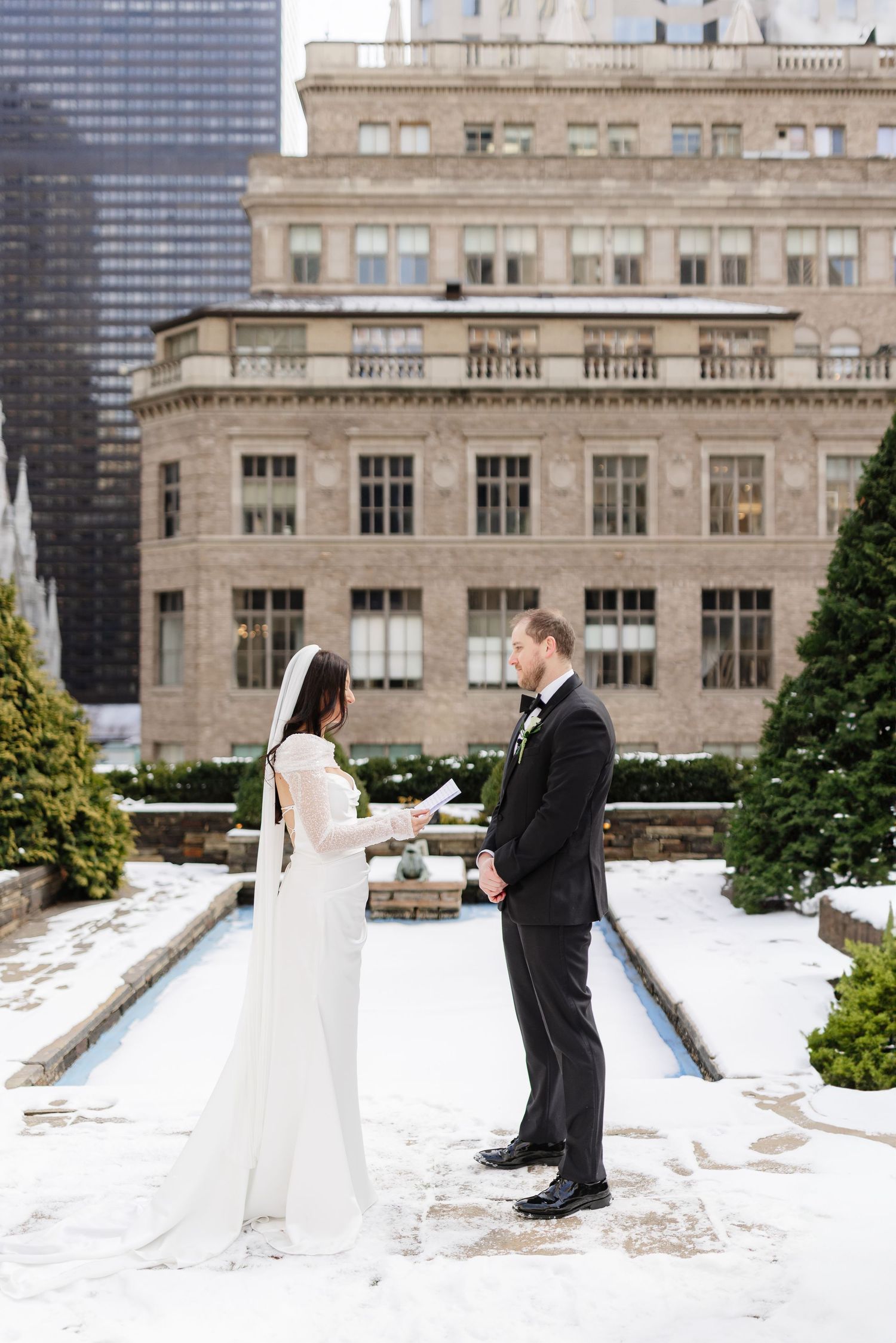 Bride and groom exchange vows in snowy Bryant Park, NYC, with classic beige building backdrop in winter.