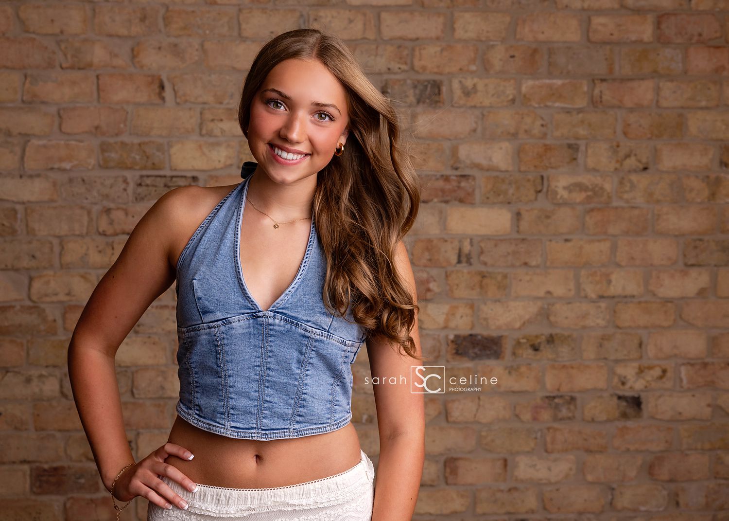 Smiling teenage girl in denim crop top and white skirt posing against a brick wall backdrop.