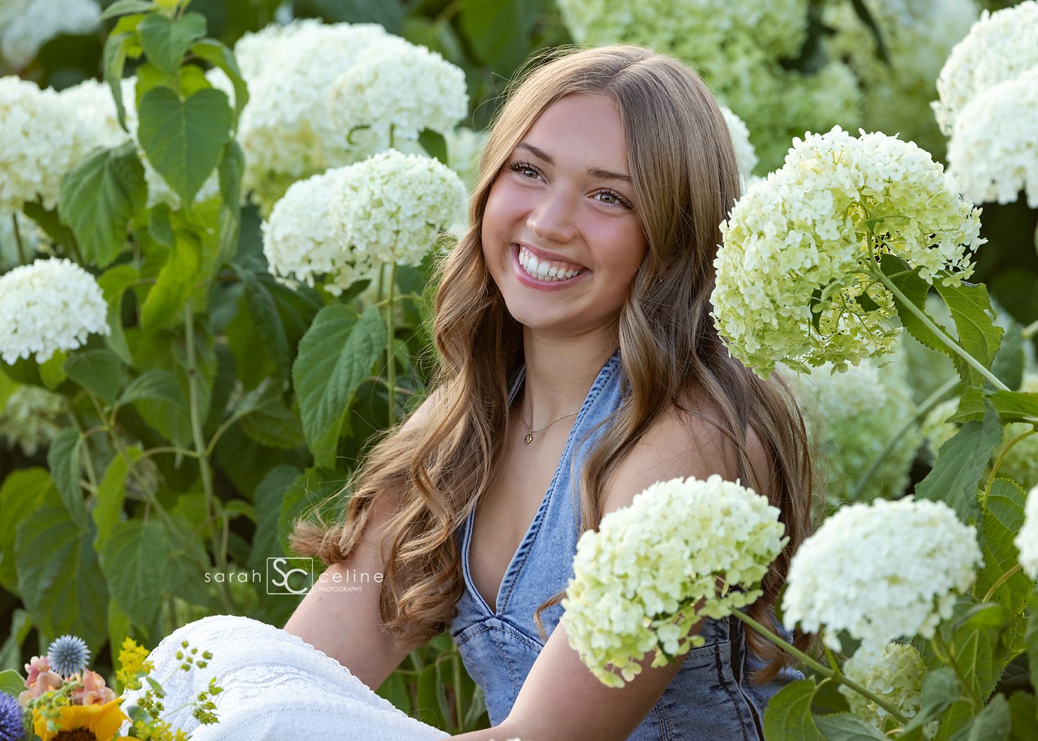 Smiling young woman holding white hydrangeas surrounded by lush blooming hydrangea bushes outdoors.