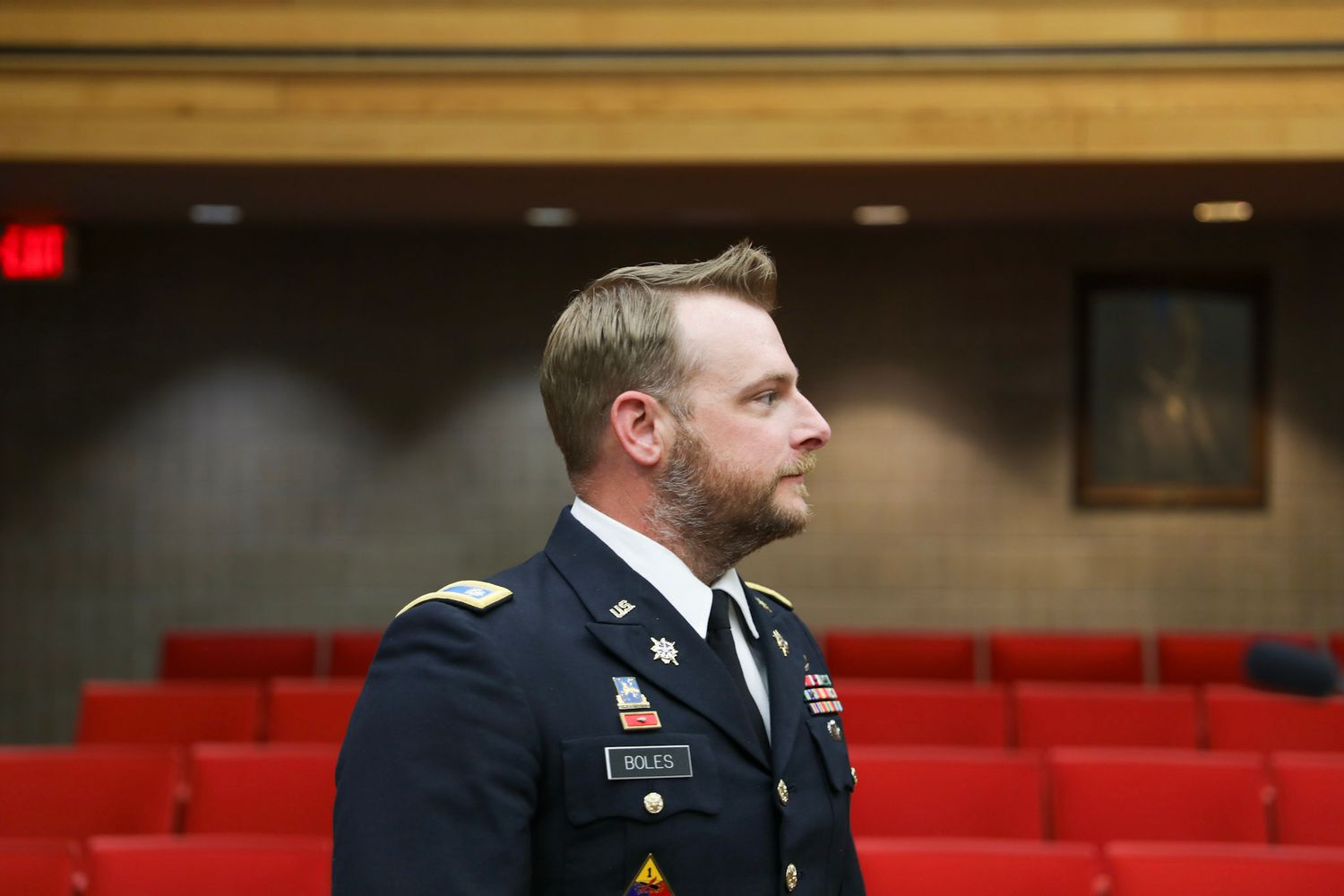 Military officer in dress uniform stands in a red-seated auditorium, wearing medals and insignia.