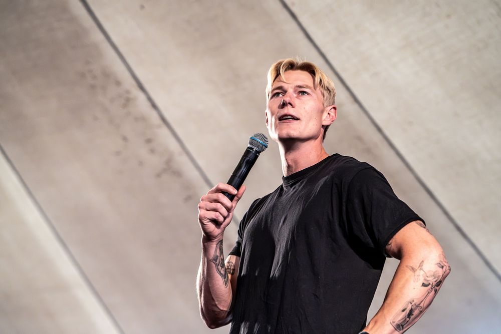 Tattooed male performer in black t-shirt holds microphone on outdoor stage against concrete wall backdrop.