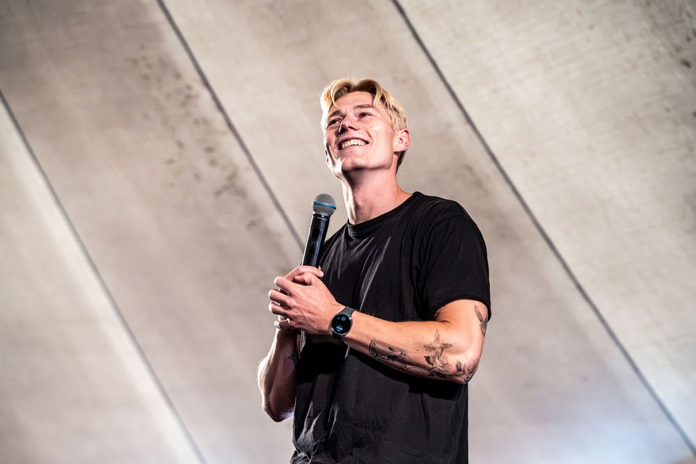 Young man in black t-shirt smiling while holding microphone on stage with dramatic lighting.