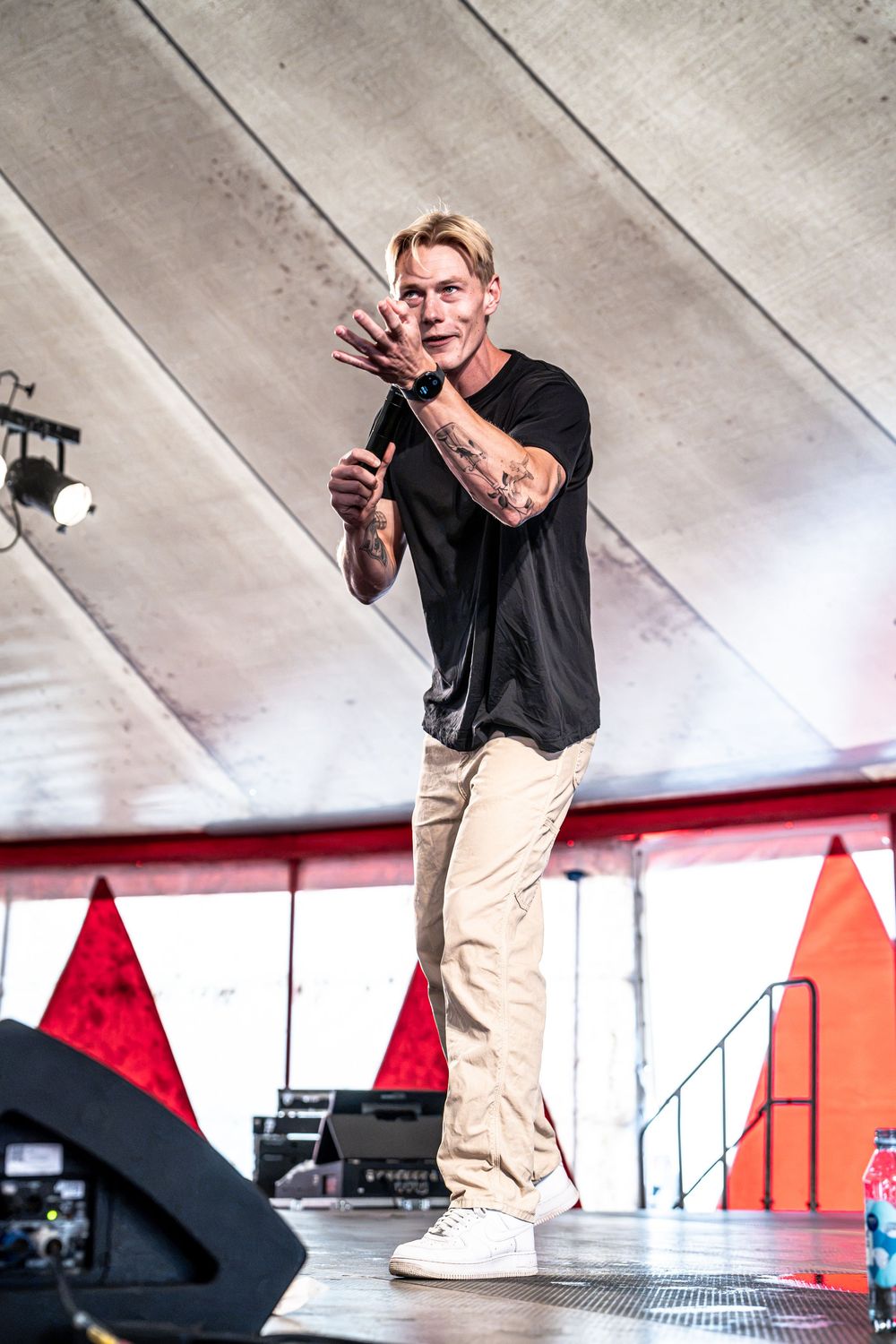 Blond male performer in black tee and khaki pants gesturing on outdoor stage with tattoos visible.