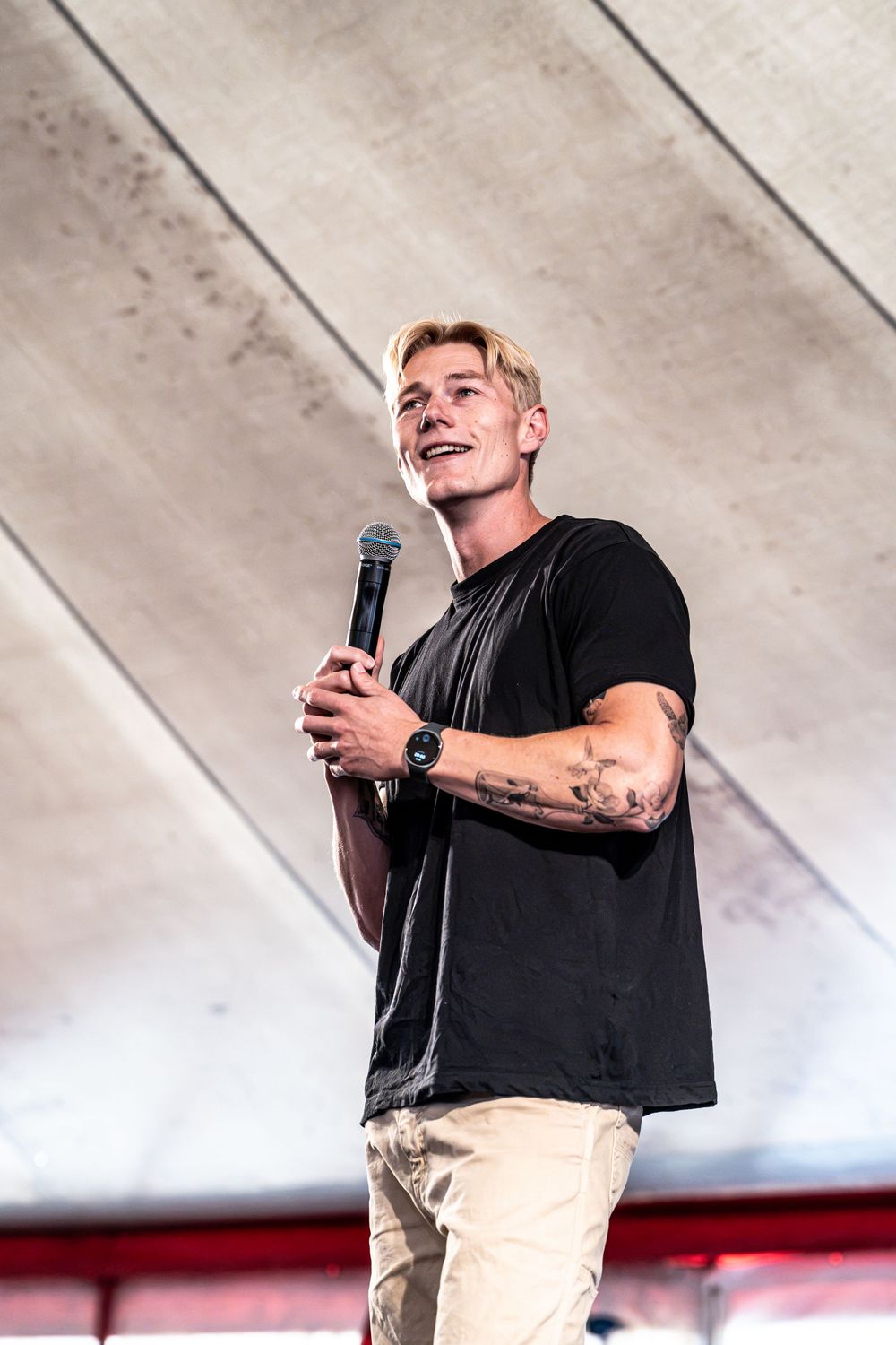 Young blond man with tattoos holding a microphone on stage, wearing a black t-shirt and khaki pants.