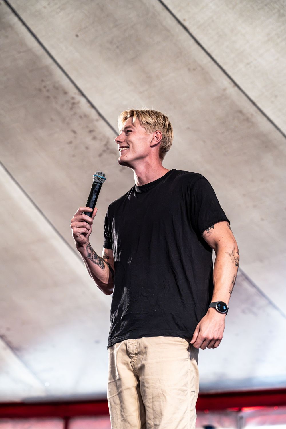 Blond male performer smiling on stage holding microphone, wearing black t-shirt and khaki pants with tattoos visible.
