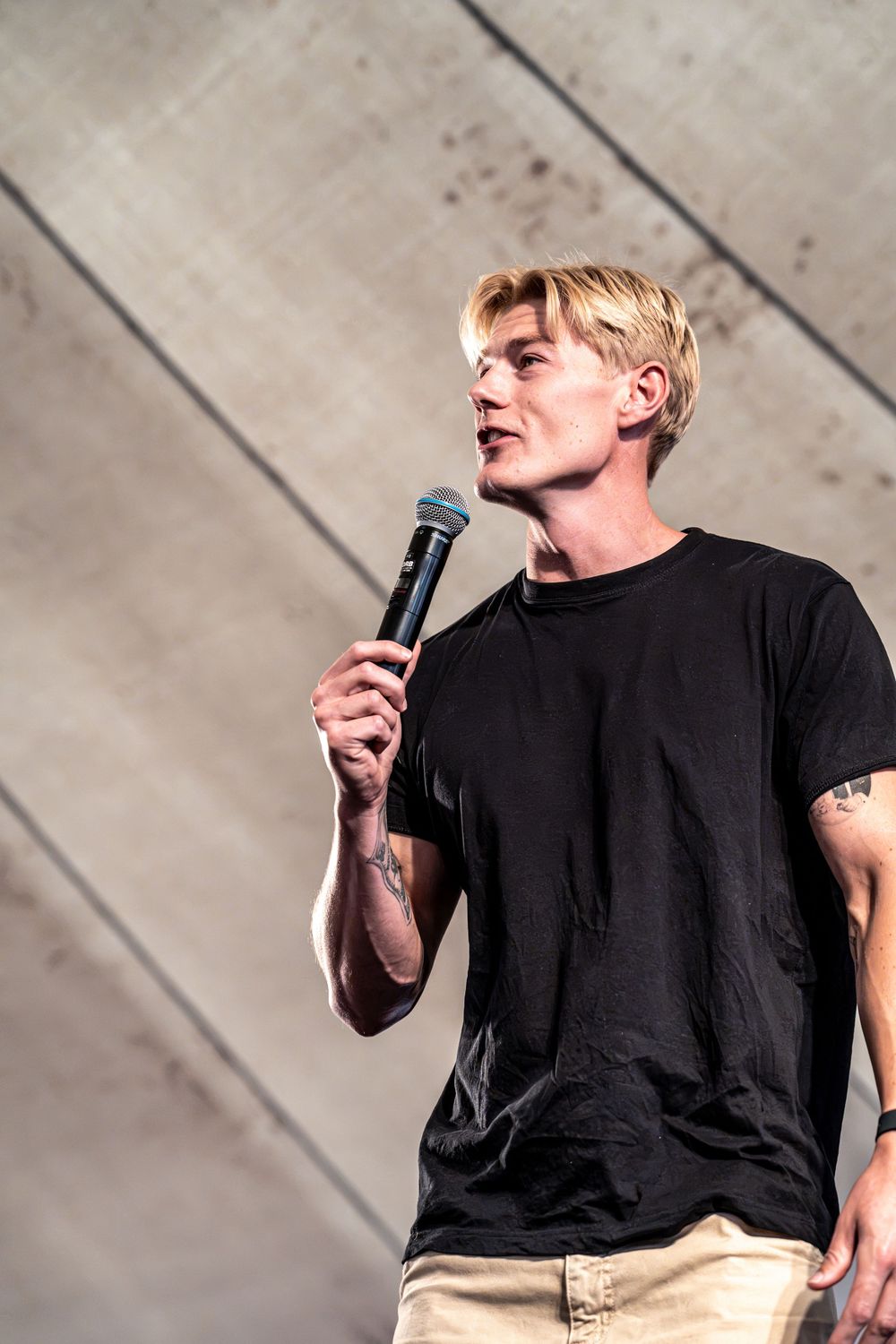 Young blonde male performer in black t-shirt holding microphone on stage with concrete wall backdrop.
