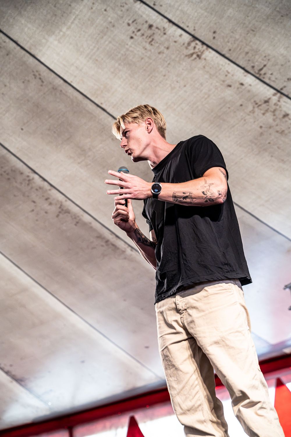Blond male performer with tattoos singing into microphone on stage wearing black t-shirt and khaki cargo pants.