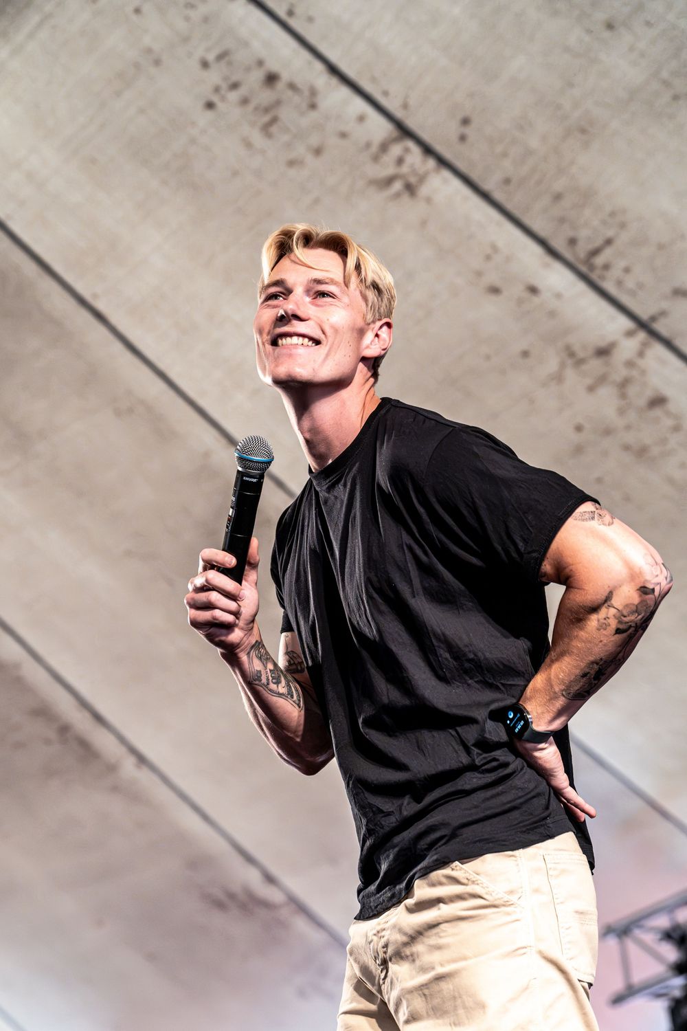 Tattooed blond man in black t-shirt smiling on stage holding a microphone with hand on hip.