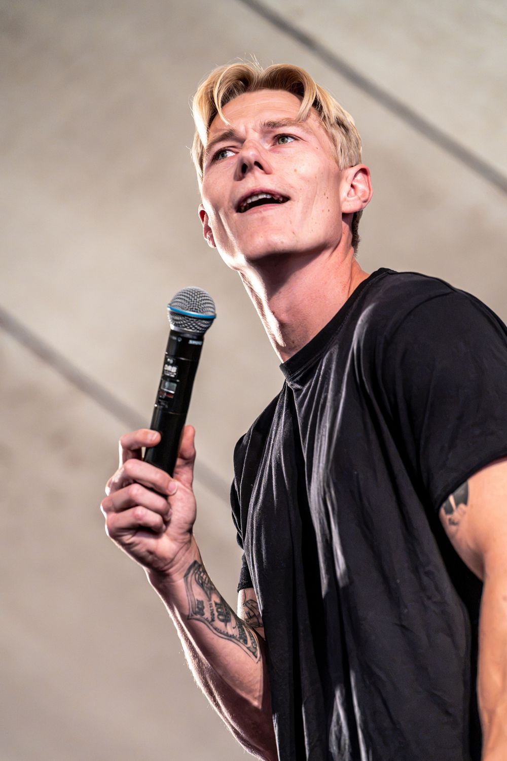 Blonde male singer with tattoos performing on stage holding a Shure microphone wearing a black t-shirt.